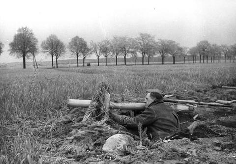 Period photograph: a man lying in a foxhole in a field with a long anti-tank tube, trees on the horizon