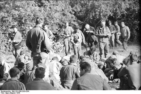 Period photograph: a crowd of men sitting on the ground among trees, guards standing nearby