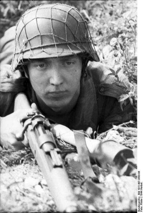 Period photograph: a young soldier in a netted helmet aiming a rifle from a foxhole