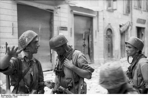 Period photograph: paratroopers in helmets talking in a street of damaged houses
