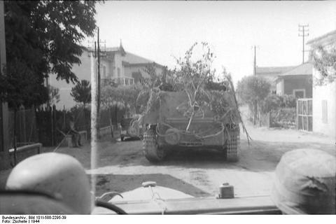 Period photograph: a half-track covered with branches seen over the helmets of soldiers in a car
