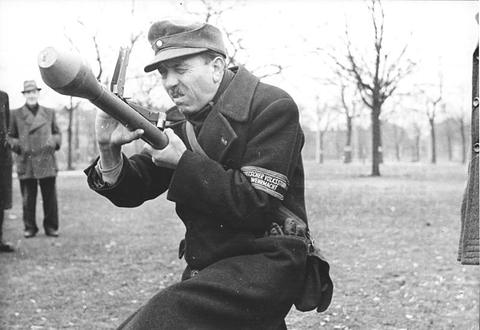 Period photograph: an older man in a cap and coat with an armband, kneeling and aiming a Panzerfaust