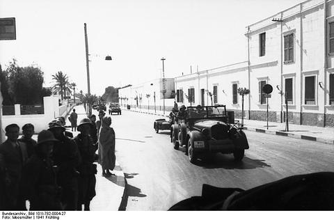 Period photograph: a car and soldiers on a wide street lined with white buildings