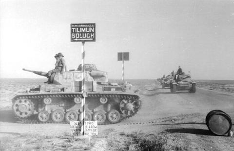 Period photograph: a tank beside a road sign, more tanks behind on a desert road