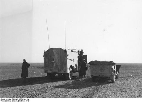 Period photograph: a large boxy armoured vehicle and a car on a desert track, a figure standing nearby