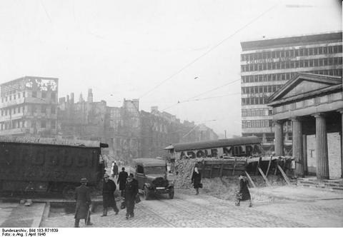 Period photograph: a timber tank barrier across a city square, pedestrians and a large modern building behind