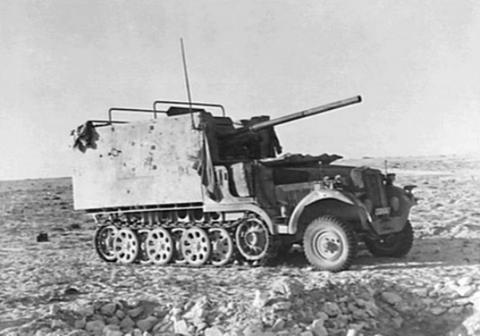 Period photograph: a half-track carrying a gun behind a pale armoured shield on stony ground