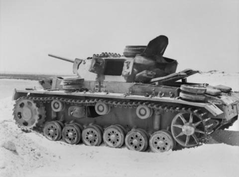 Period photograph: a damaged tank sunk in pale sand, spare wheels on its hull