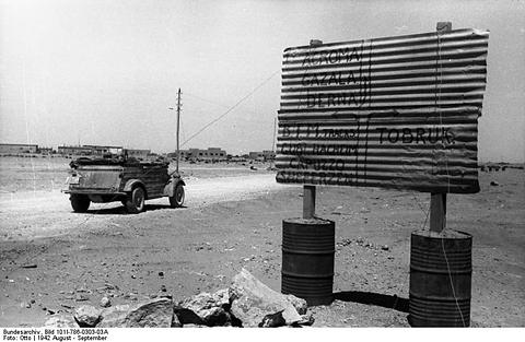 Period photograph: a small car on a road beside a large sign mounted on oil drums