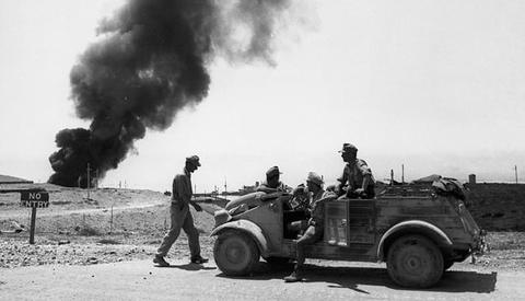 Period photograph: soldiers in an open car on a road, a tall plume of black smoke behind