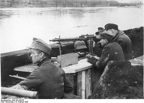 Period photograph: men in caps and greatcoats at a machine gun behind a parapet, the river behind them
