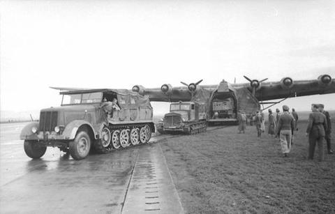 Period photograph: half-tracks in front of a very large aircraft with its nose doors open