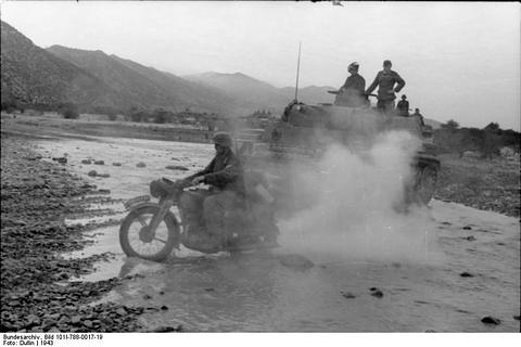 Period photograph: a motorcycle and a tank splashing through a shallow river