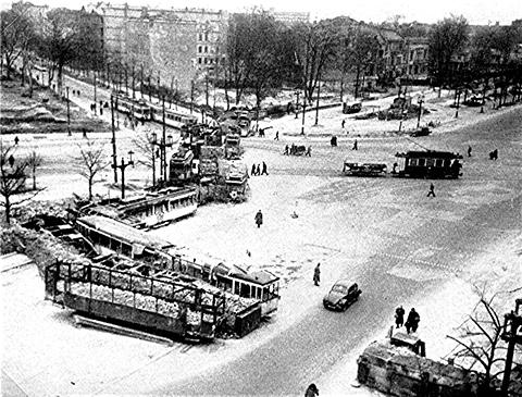 Period photograph: a city square seen from above, with tram cars pushed together as barricades