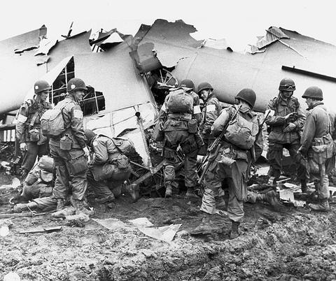Period photograph: American paratroopers in helmets gathered around the torn fuselage of a glider on muddy ground