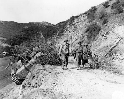 Period photograph: soldiers walking along a mountain road past a wrecked tank