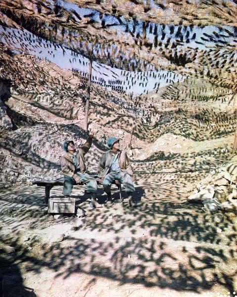 Period photograph (wartime colour): a hillside gun position hidden under netting, two American soldiers at its entrance