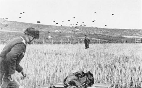 Period photograph: a paratrooper in a jump smock in a grain field, parachutes in the sky behind