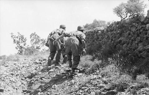 Period photograph: two paratroopers seen from behind, in smocks, webbing and helmets, moving along a stone wall