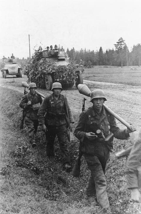 Period photograph: soldiers in helmets and smocks walking along a muddy track, a half-track behind them