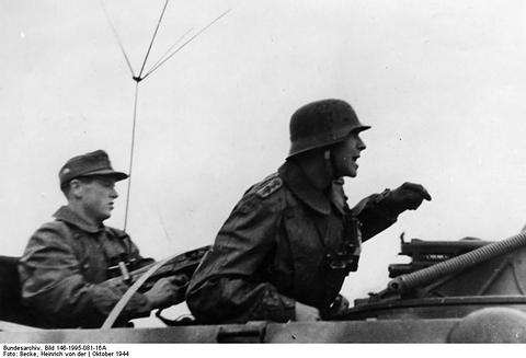 Period photograph: two soldiers in steel helmets in the open top of a half-track, one pointing, a radio antenna above them