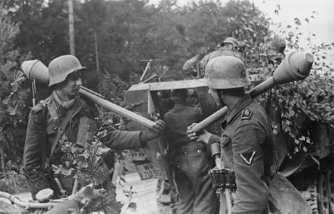 Period photograph: soldiers in steel helmets advancing through trees, carrying Panzerfausts over their shoulders