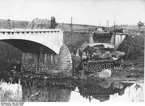Period photograph: a tank lying on the riverbank below the broken span of a road bridge, figures on the bank above