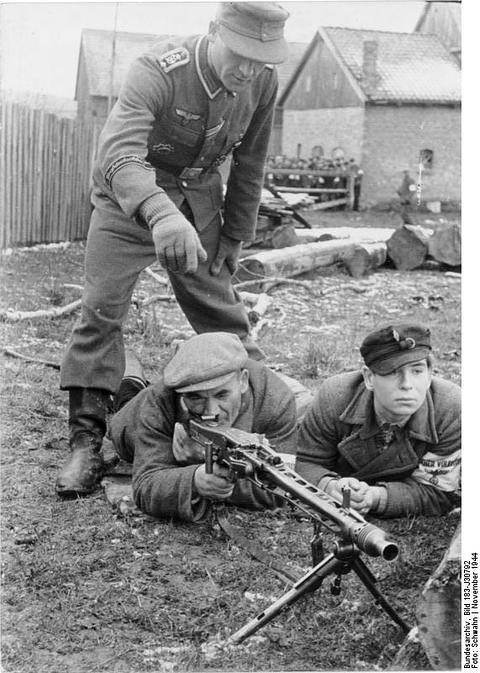 Period photograph: a soldier standing over two men lying behind a machine gun on a bipod, one with an armband
