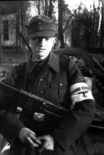 Period photograph: a young man in a field cap and coat holding a submachine gun, an armband on his left arm