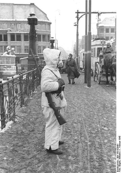 Period photograph: a sentry in a white hooded suit standing on a cobbled bridge, horse-drawn carts behind