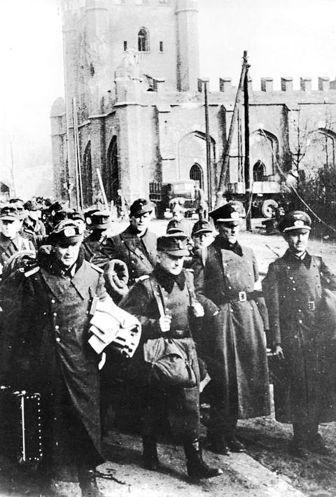 Period photograph: a group of officers in peaked caps and greatcoats standing in a street in front of a brick gate building