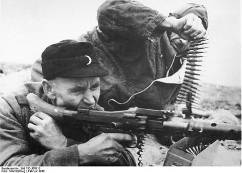 Period photograph: close view of a man in a dark cap aiming a machine gun, an ammunition belt held up beside him