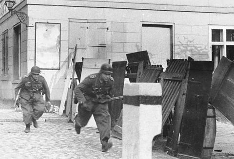 Period photograph: two soldiers running bent over past wooden barricades in a town street