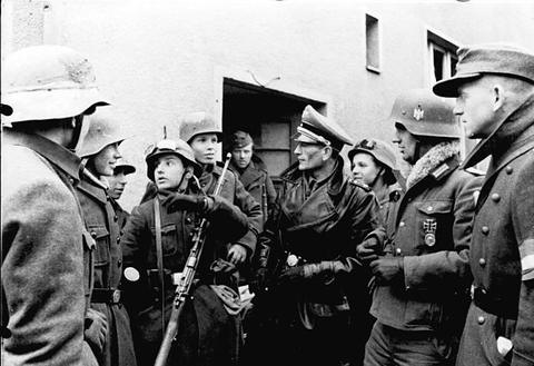 Period photograph: a group of boys in steel helmets holding rifles, gathered around a man in a peaked cap