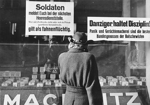 Period photograph: a woman in a coat seen from behind, reading printed notices in a shop window