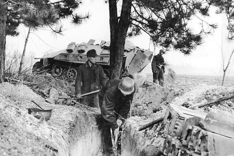 Period photograph: young men digging in sandy ground under a tree, a half-track in the background