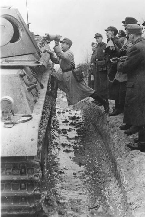 Period photograph: a tank driving past a trench, men crouching in the trench and others watching beside it