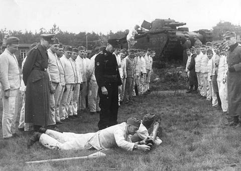 Period photograph: a man lying on the grass aiming a rocket launcher, a line of men watching and a tank behind