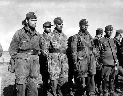 Period photograph: a line of officers in caps, several in camouflage smocks and trousers, standing in the open
