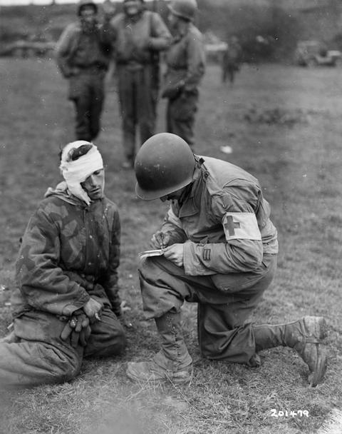 Period photograph: a kneeling prisoner with a bandaged head in a hooded smock, an American soldier with a Red Cross armband writing beside him