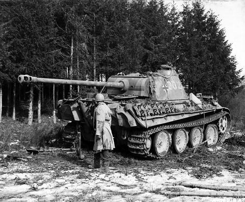 Period photograph: a Panther tank in camouflage paint in front of fir trees, a soldier in a coat standing beside it