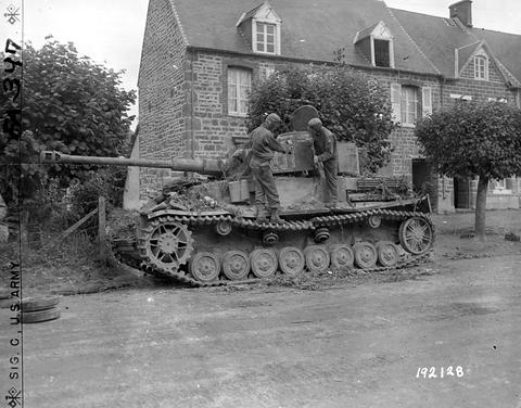 Period photograph: a camouflaged Panzer IV with side skirts in front of a stone house, two soldiers on the hull