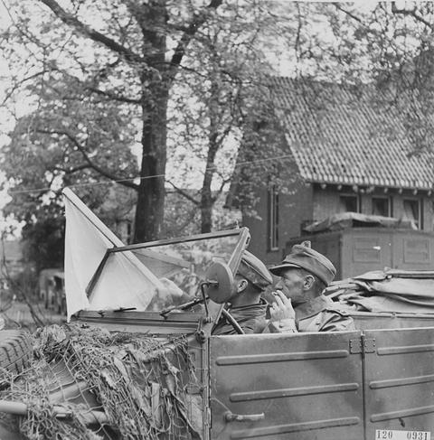 Period photograph: two officers in caps seated in an open car with a white flag, netting and foliage over its front