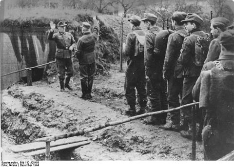 Period photograph: a file of men in caps and coats with rifles standing beside a bunker covered with earth