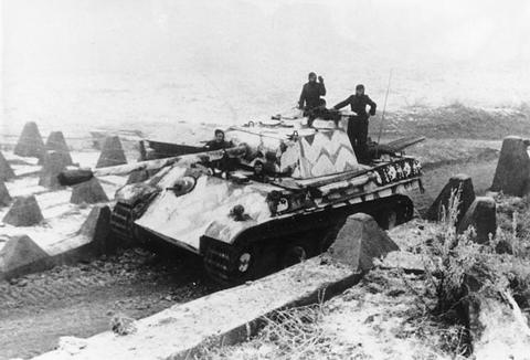 Period photograph: a camouflage-painted Panther tank with men on the hull driving between concrete tank obstacles