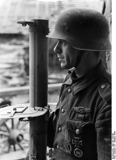 Period photograph: a soldier in a steel helmet seen in profile, the tube of a rocket launcher on his shoulder, decorations on his tunic